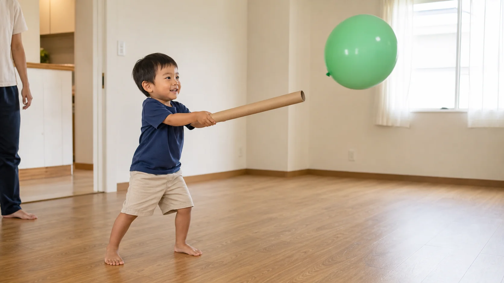A toddler batting a large balloon with a cardboard wrapping-paper tube in a cleared living room while a grown-up watches nearby.