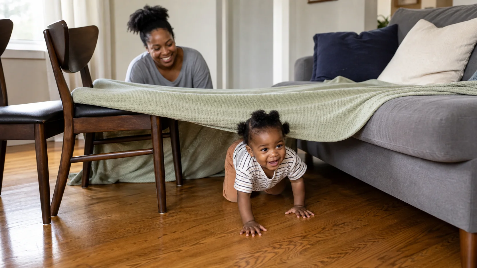 A toddler crawling into a blanket fort stretched between a couch and chairs while a grown-up stays nearby.