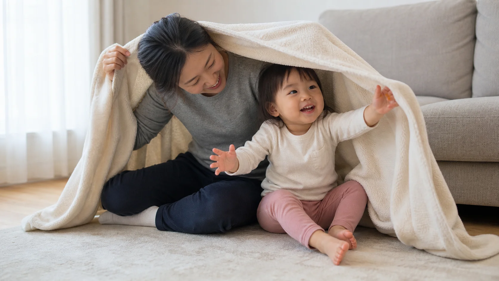 A toddler and grown-up sitting together on a couch as a loose blanket lifts for a quick hide-and-reveal game.