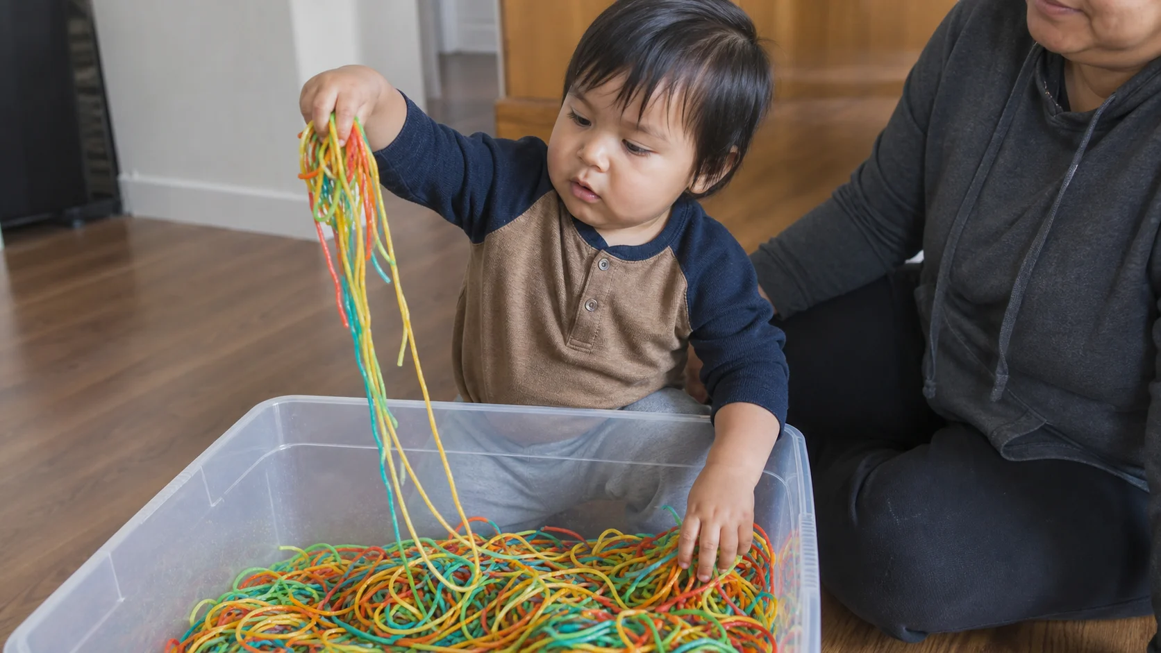 A toddler sitting beside a large tub of cooked coloured spaghetti, lifting one slippery handful while a grown-up stays close nearby.