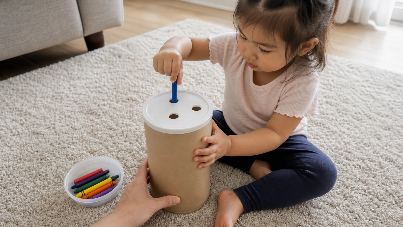 A toddler pushing a crayon through a hole in the lid of an oats container while other crayons sit in a small bowl.