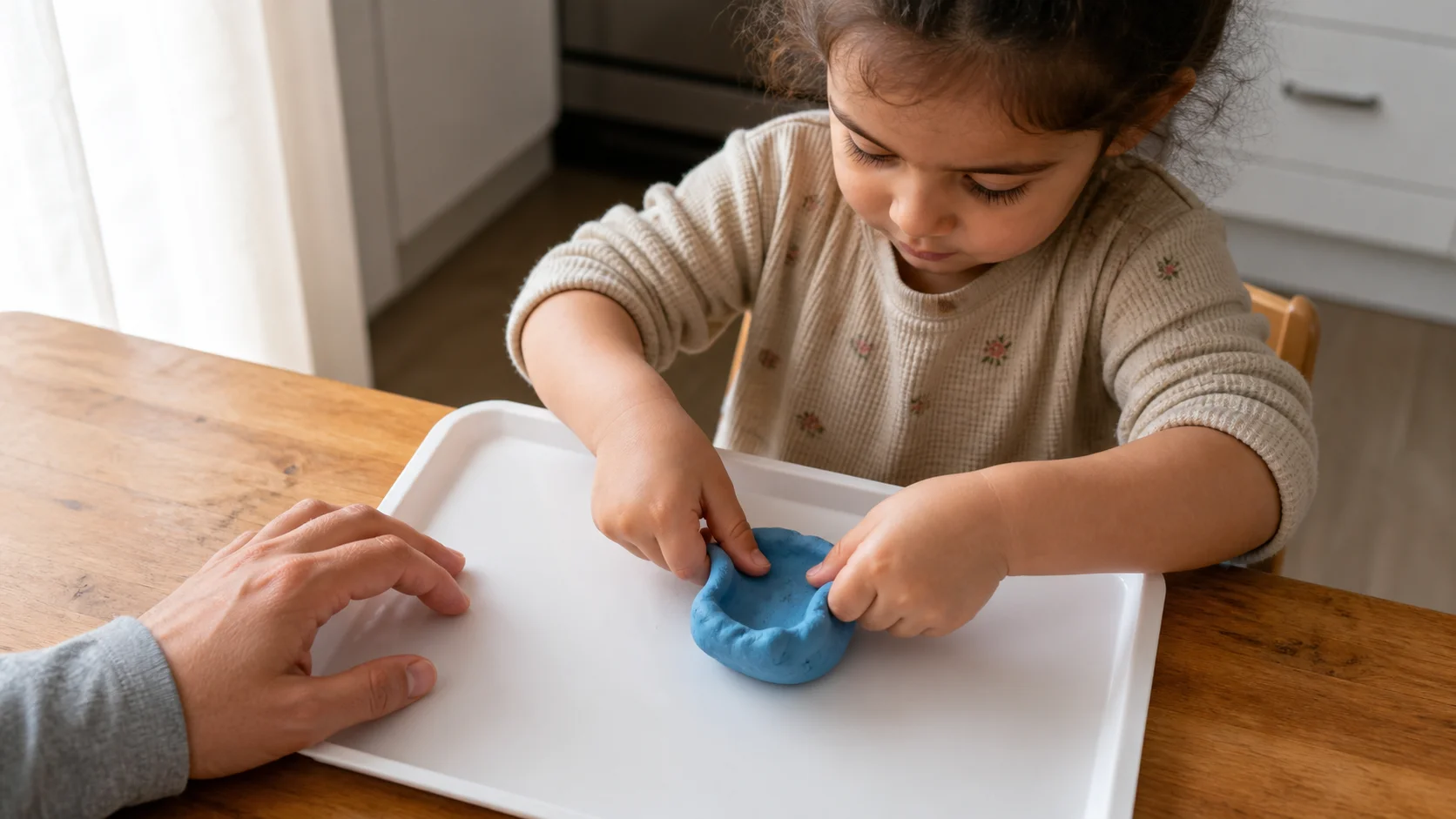 Toddler pressing thumbs into a dough ball on a tray while a grown-up steadies the setup.