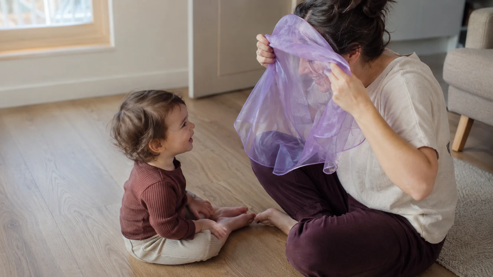 A grown-up sitting face to face with a toddler while a light scarf floats down in a simple reveal game.