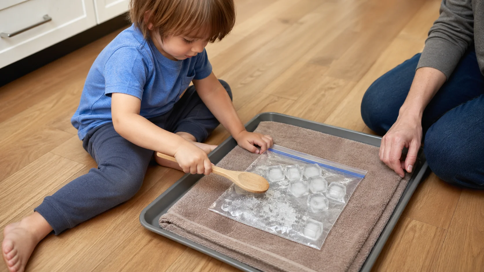A young child taps a sealed bag of ice cubes with a wooden spoon on a towel while a grown-up sits nearby.