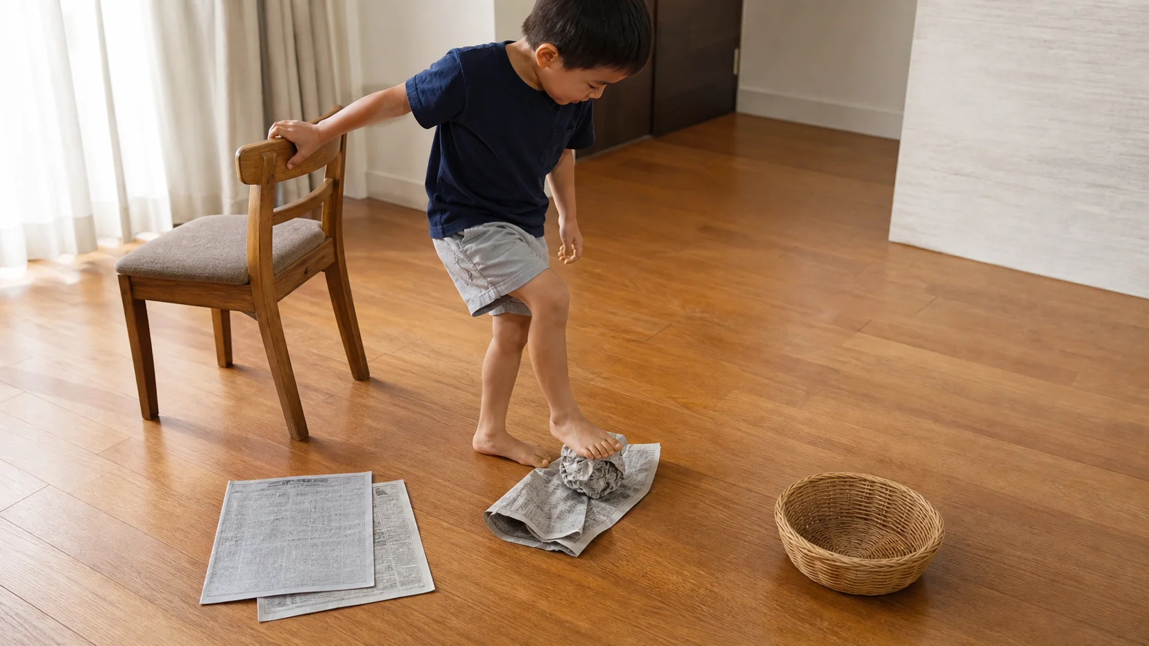 Preschool child balancing beside a chair while crumpling newspaper with one foot near a basket.