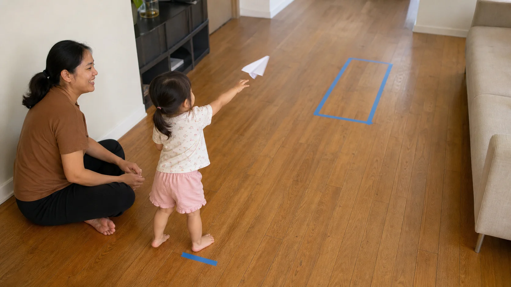 A toddler launching a simple paper plane toward a nearby landing strip while a grown-up sits beside the start spot.