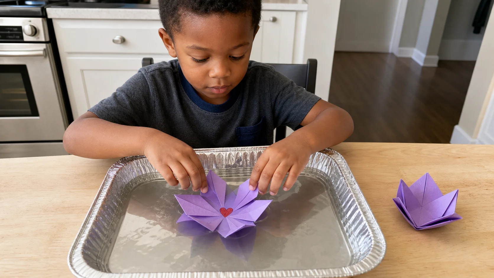 Preschool child placing a folded paper flower onto a shallow foil pan of water while a grown-up sits nearby.