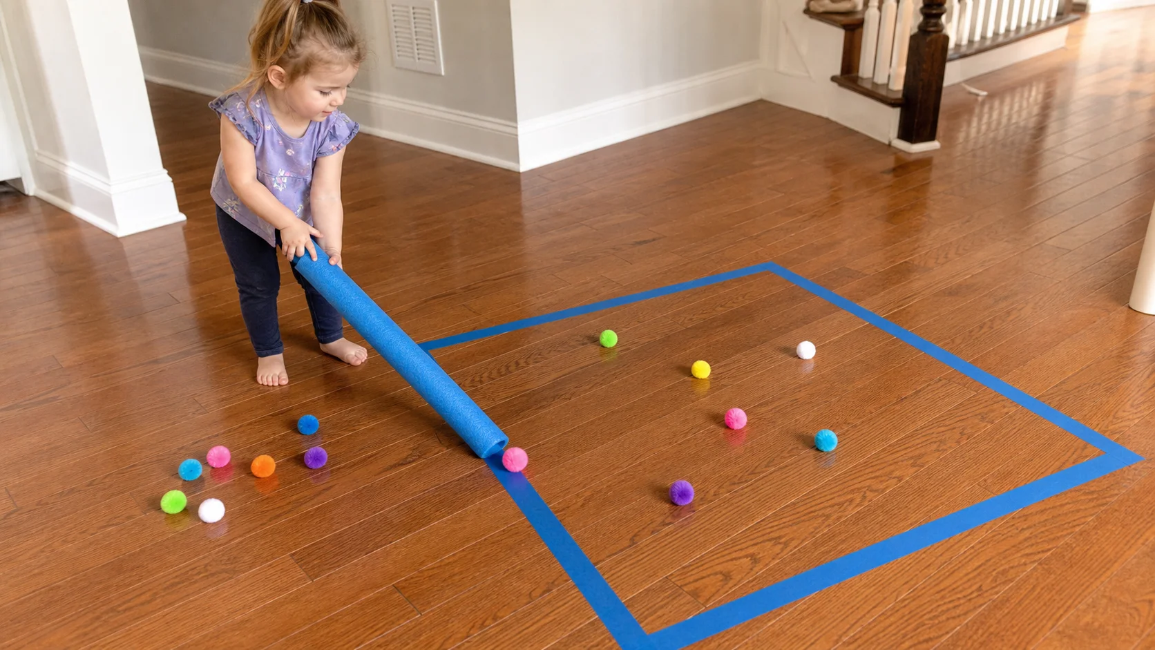 A toddler kneels beside a taped floor square and pushes a bright pom-pom toward it with a pool noodle.