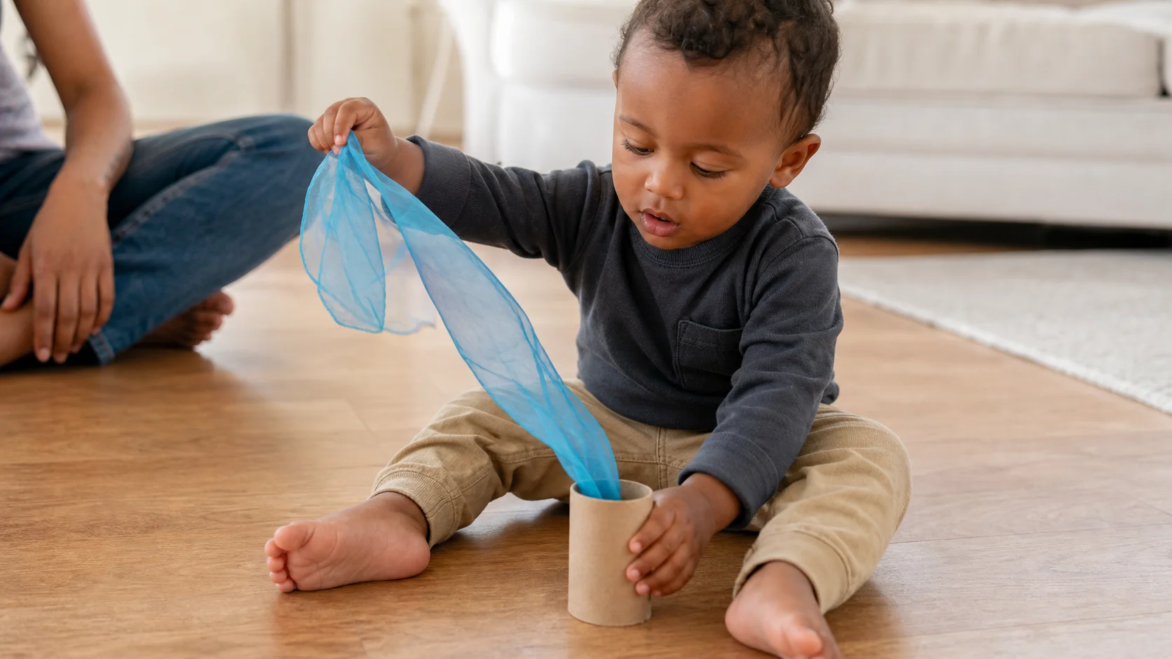 A toddler sitting on the floor pushing a colorful scarf into a cardboard tube while a grown-up stays nearby.