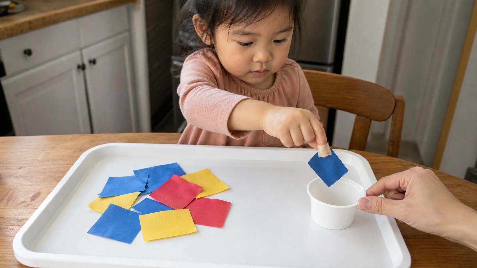 Child using a loose sticky tape loop on one fingertip to pick up a large paper scrap beside a small cup on a tray.