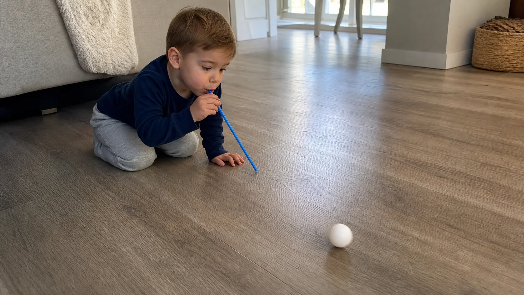 A young child leaning over a straw and blowing a lightweight ball across a short clear floor lane while a grown-up sits nearby.