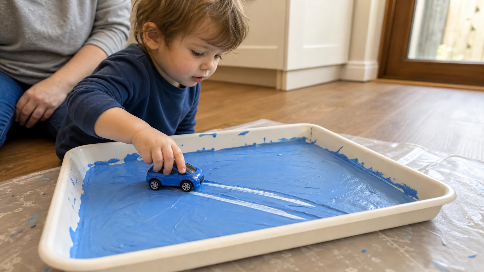 Toddler sitting beside a shallow tray of washable paint and pushing a familiar toy car through it to make clear tracks