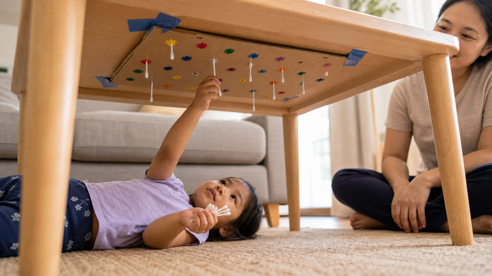 A child lies under a low table and pushes Q-tips into a cardboard star board taped overhead.