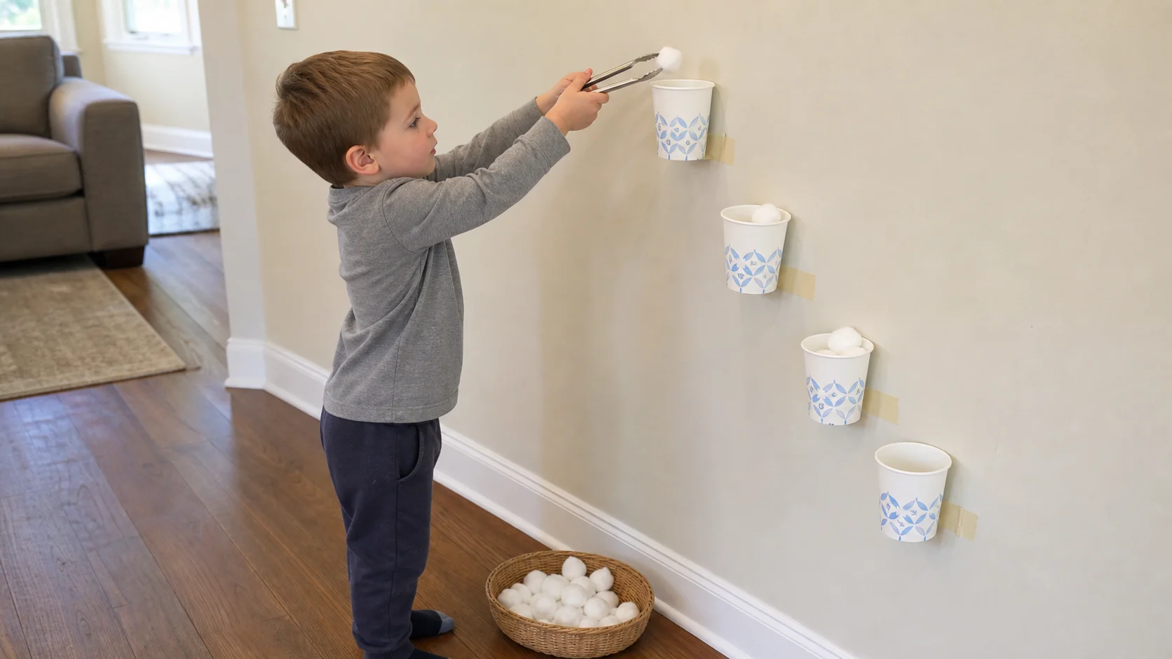 A child stands by a wall and drops a cotton ball from tongs into a paper cup target.